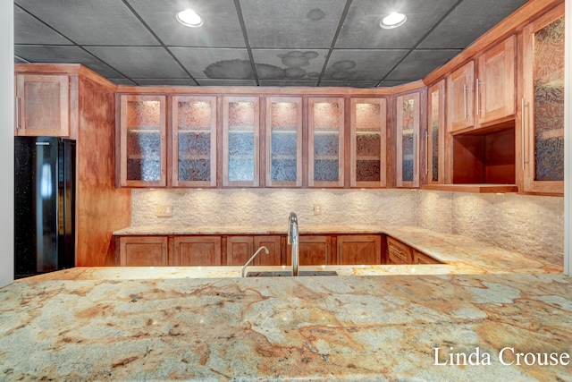 kitchen featuring tasteful backsplash, glass insert cabinets, freestanding refrigerator, a sink, and light stone countertops