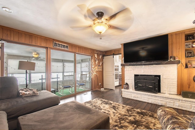 living room featuring ceiling fan, wooden walls, dark hardwood / wood-style floors, a stone fireplace, and a barn door