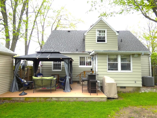 rear view of house with a lawn, cooling unit, a gazebo, a wooden deck, and roof with shingles
