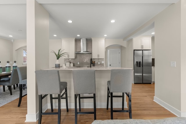 kitchen featuring stainless steel refrigerator with ice dispenser, wall chimney exhaust hood, kitchen peninsula, and white cabinets