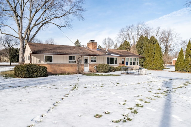 snow covered back of property featuring board and batten siding, brick siding, and a chimney