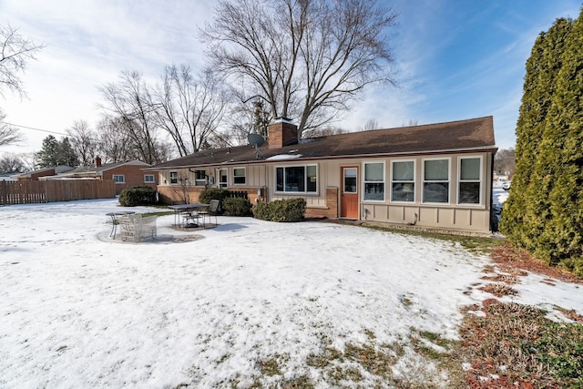 snow covered house featuring board and batten siding, brick siding, fence, and a chimney