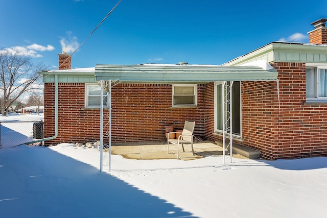 snow covered house with entry steps, a patio, a chimney, fence, and brick siding