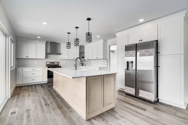 kitchen with a center island with sink, white cabinets, wall chimney exhaust hood, appliances with stainless steel finishes, and decorative light fixtures