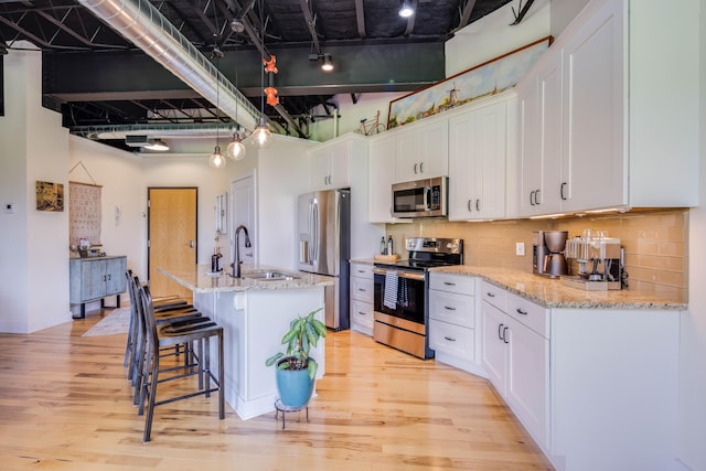 kitchen featuring white cabinetry, a towering ceiling, stainless steel appliances, light stone countertops, and a kitchen island with sink