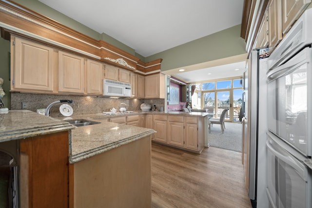 kitchen featuring light stone counters, a peninsula, white appliances, a sink, and light brown cabinetry