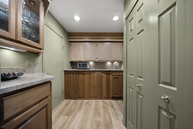 kitchen with light wood finished floors, glass insert cabinets, light stone counters, backsplash, and a sink