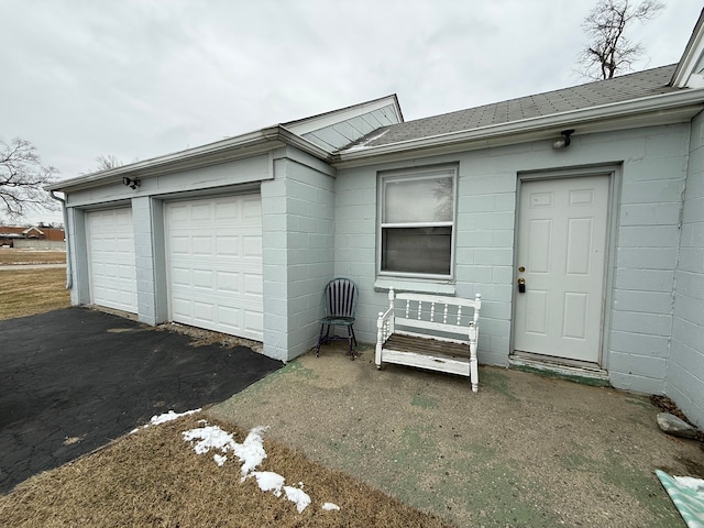 view of exterior entry featuring an attached garage, concrete block siding, aphalt driveway, and roof with shingles