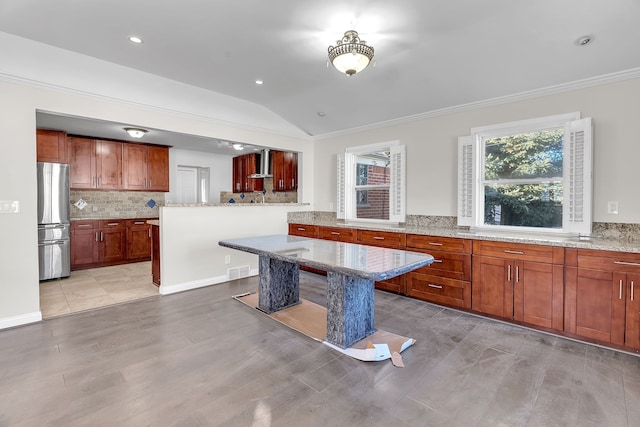 kitchen with a kitchen island, light stone counters, freestanding refrigerator, wall chimney range hood, and backsplash