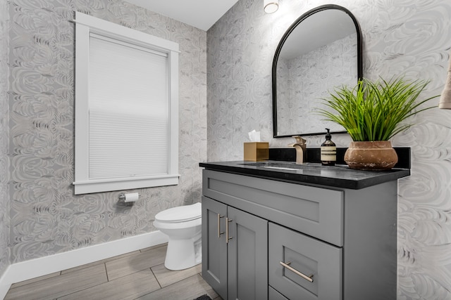 half bathroom with toilet, baseboards, vanity, and wallpapered walls