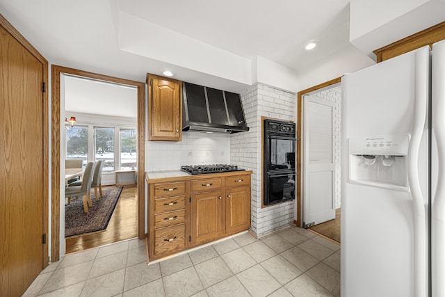 kitchen featuring white refrigerator with ice dispenser, tile countertops, black double oven, wall chimney exhaust hood, and stainless steel gas cooktop