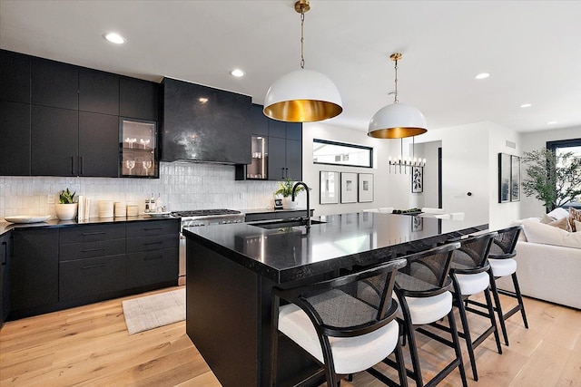 kitchen featuring dark countertops, pendant lighting, wall chimney range hood, and an island with sink