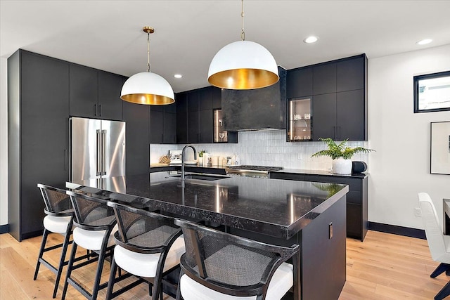 kitchen with decorative light fixtures, dark countertops, high quality fridge, a sink, and an island with sink