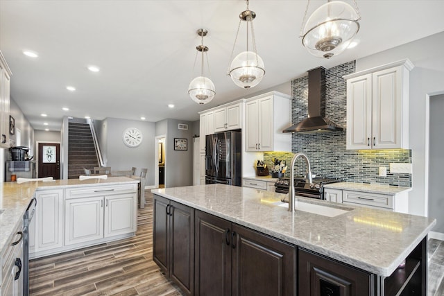 kitchen featuring decorative light fixtures, white cabinetry, an island with sink, black fridge with ice dispenser, and wall chimney range hood