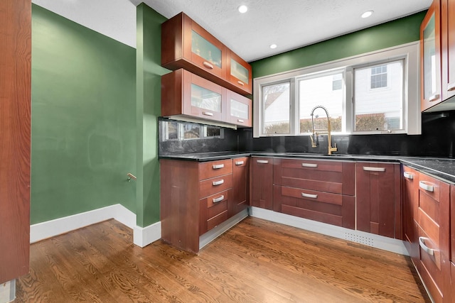 kitchen featuring baseboards, a sink, decorative backsplash, dark wood finished floors, and glass insert cabinets