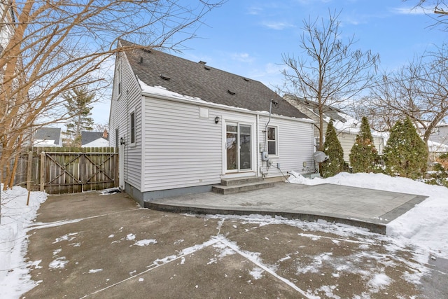 snow covered house with a patio, a shingled roof, entry steps, a gate, and fence