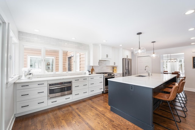 kitchen featuring stainless steel appliances, hanging light fixtures, a kitchen island with sink, a sink, and white cabinetry