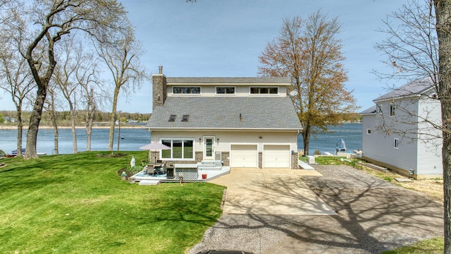 back of house featuring roof with shingles, a chimney, concrete driveway, a lawn, and a water view