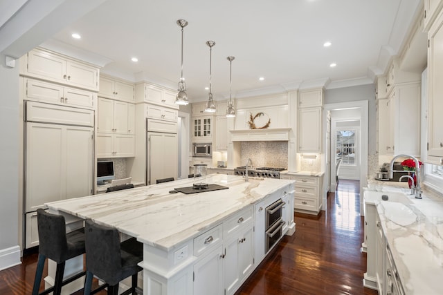 kitchen featuring built in appliances, light stone counters, decorative light fixtures, ornamental molding, and a large island with sink