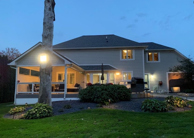 back of property at dusk with roof with shingles, a lawn, and a patio