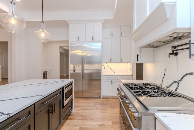 kitchen with custom exhaust hood, white cabinetry, hanging light fixtures, and built in appliances