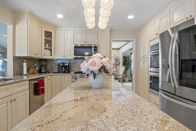 kitchen with cream cabinetry, hanging light fixtures, appliances with stainless steel finishes, glass insert cabinets, and light stone countertops