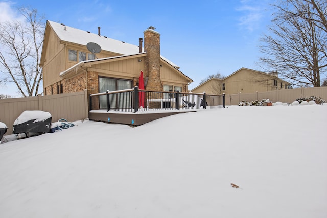 snow covered house featuring a wooden deck, a chimney, fence, and brick siding