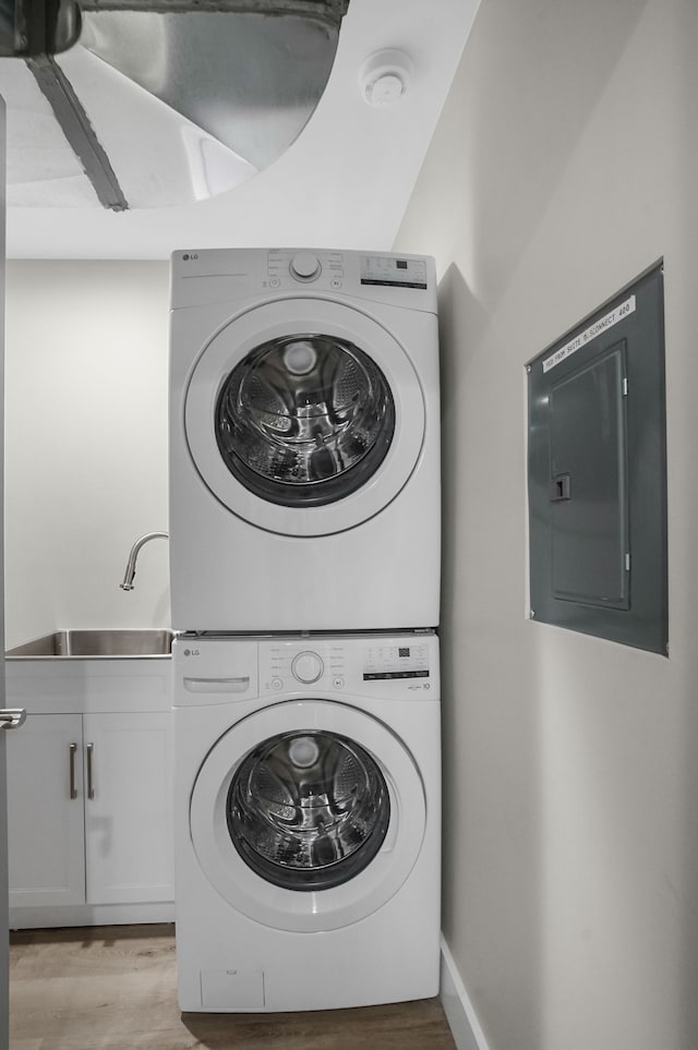 clothes washing area with a sink, stacked washing maching and dryer, cabinet space, electric panel, and light wood finished floors