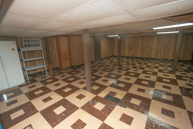 finished below grade area featuring wood walls, dark floors, a drop ceiling, and freestanding refrigerator