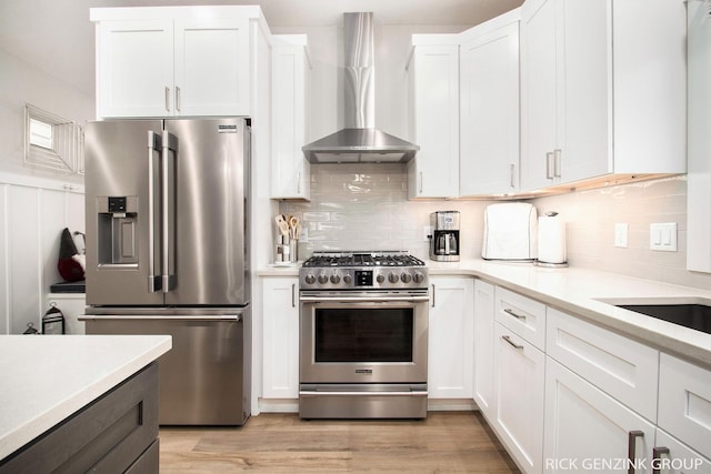 kitchen featuring wall chimney exhaust hood, tasteful backsplash, light hardwood / wood-style flooring, stainless steel appliances, and white cabinets