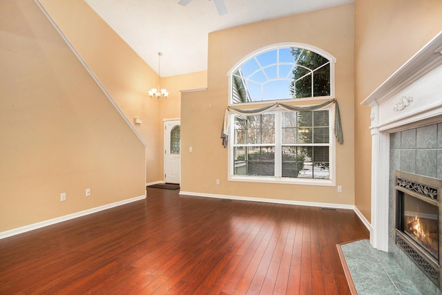 unfurnished living room with ceiling fan with notable chandelier, a tiled fireplace, dark wood-type flooring, and a high ceiling