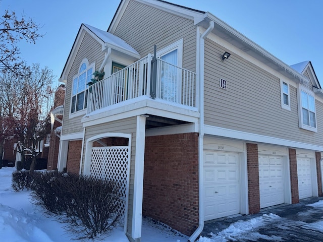 snow covered property with brick siding, an attached garage, and a balcony