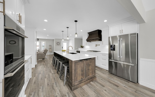 kitchen with a center island with sink, white cabinets, custom exhaust hood, stainless steel appliances, and a sink