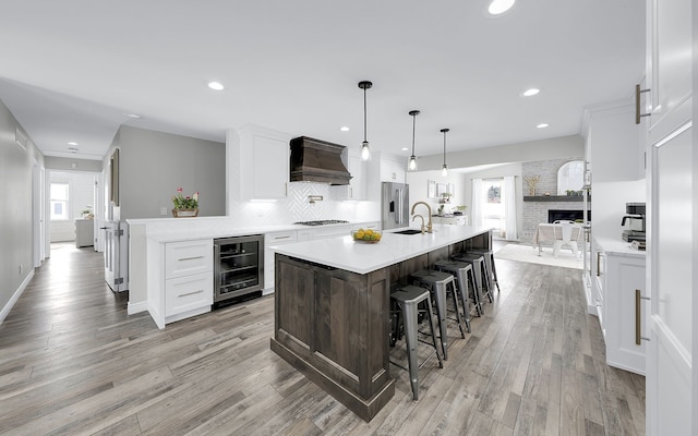 kitchen with white cabinets, an island with sink, custom range hood, wine cooler, and light countertops