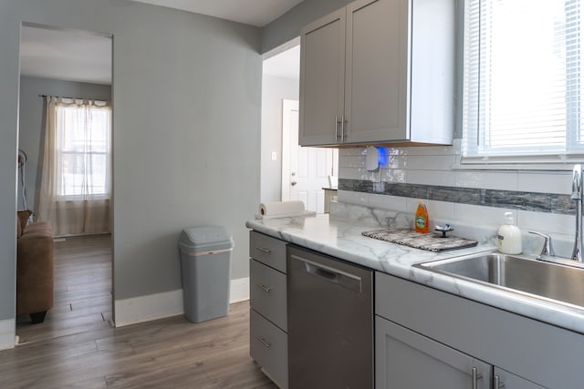 kitchen with wood finished floors, stainless steel dishwasher, a sink, backsplash, and gray cabinetry