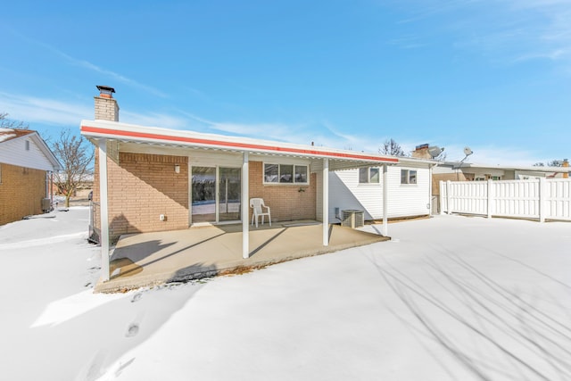 snow covered property featuring brick siding, a patio, a chimney, fence, and cooling unit