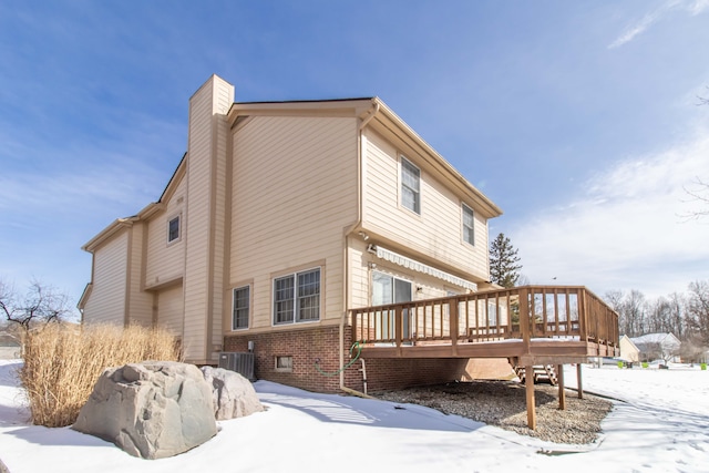 snow covered property with a deck, brick siding, a chimney, and central AC unit