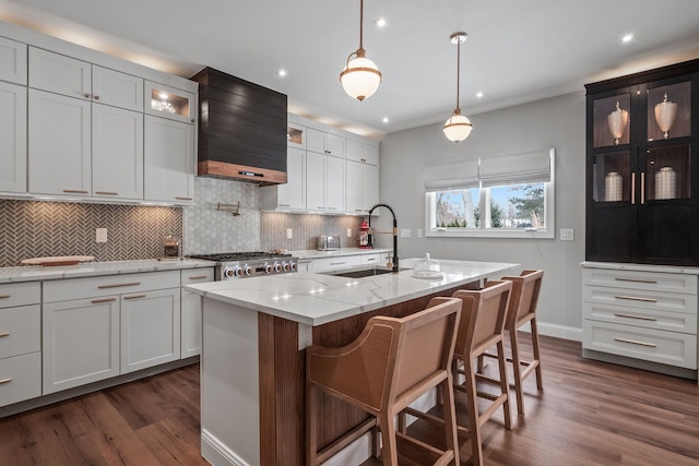 kitchen with light stone counters, a kitchen island with sink, a sink, custom exhaust hood, and glass insert cabinets