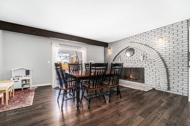 dining room featuring brick wall, dark wood finished floors, baseboards, beam ceiling, and a fireplace