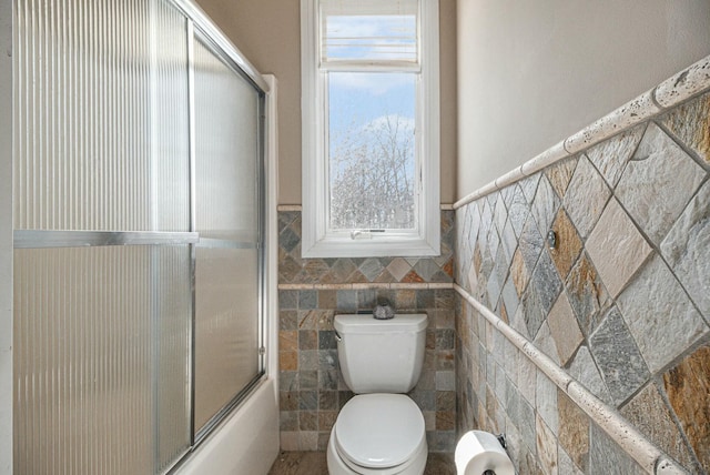 full bathroom featuring shower / bath combination with glass door, wainscoting, toilet, and tile walls