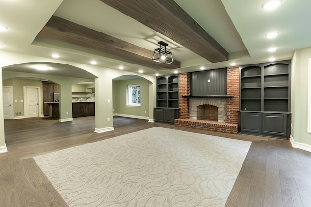 unfurnished living room featuring a raised ceiling, beamed ceiling, dark hardwood / wood-style floors, a brick fireplace, and built in features