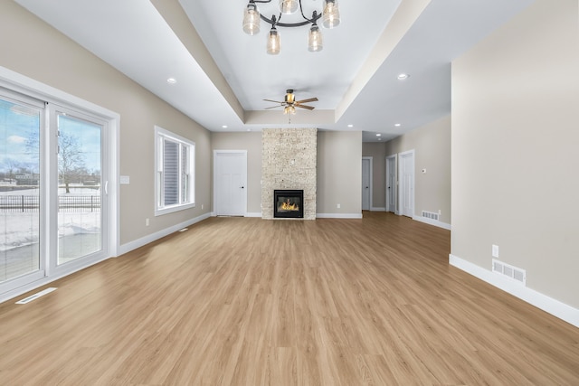 unfurnished living room featuring a raised ceiling, light wood-type flooring, ceiling fan with notable chandelier, and a stone fireplace