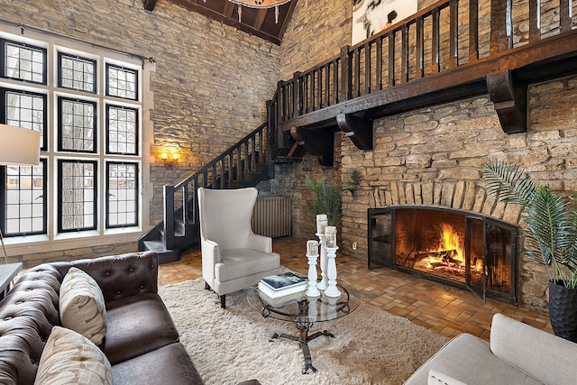 living area featuring a warm lit fireplace, brick floor, a high ceiling, stairway, and beam ceiling