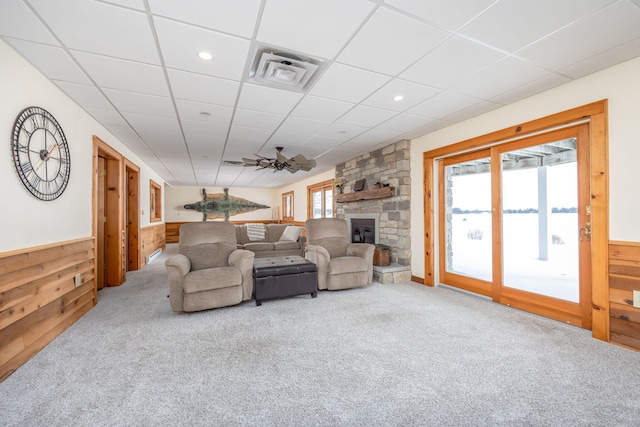 carpeted living room with a paneled ceiling, ceiling fan, wood walls, and a stone fireplace