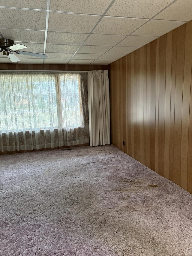 carpeted empty room featuring a paneled ceiling, wooden walls, and ceiling fan