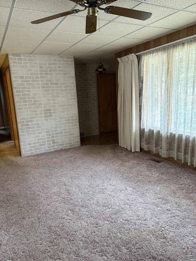 carpeted empty room featuring ceiling fan, brick wall, and a drop ceiling