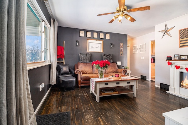 living room featuring dark wood-style flooring, visible vents, a ceiling fan, baseboards, and a lit fireplace