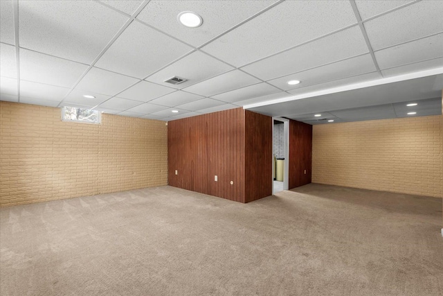 basement with a paneled ceiling, visible vents, light carpet, wood walls, and brick wall