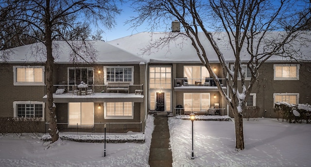 snow covered house featuring brick siding, a chimney, and a balcony