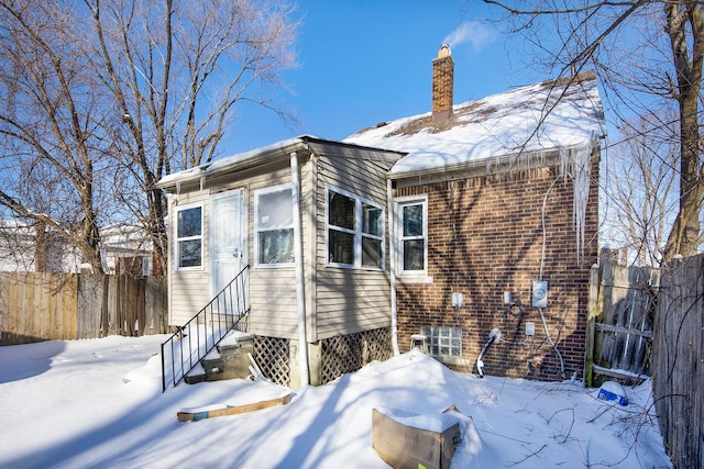 snow covered back of property featuring entry steps, brick siding, fence, and a chimney